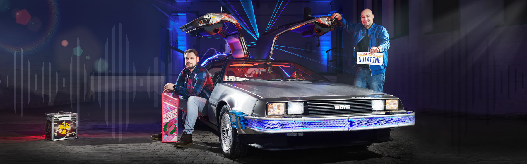 Podcast OUTATIME Header Zwei Männer posieren vor einem DeLorean mit geöffneten Flügeltüren, inspiriert von „Zurück in die Zukunft“. Einer sitzt mit einem Hoverboard, der andere hält ein Nummernschild mit der Aufschrift „OUTATIME“.