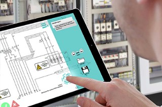 Tablet in einer Industriehalle zeigt den Plan einer Schaltanlage, eine Hand zeigt darauf.