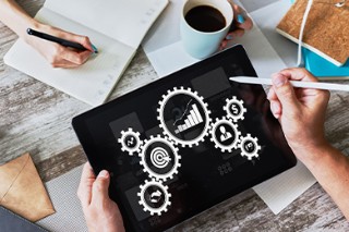 Two hands above a table. One of them is holding a tablet, the other a tablet pen. The display shows graphics for development analysis.