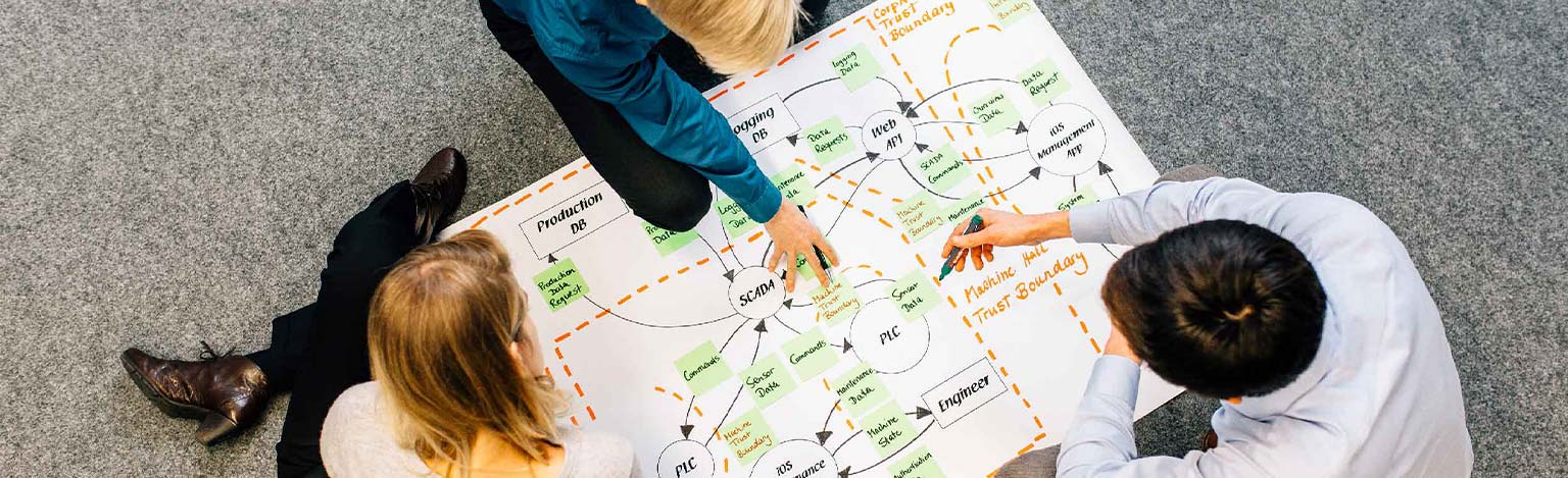 A bird's eye view of three people at a table. They develop a schematic drawing on the table.