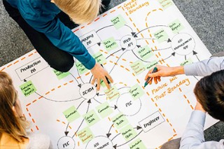 A bird's eye view of three people at a table. They develop a schematic drawing on the table.